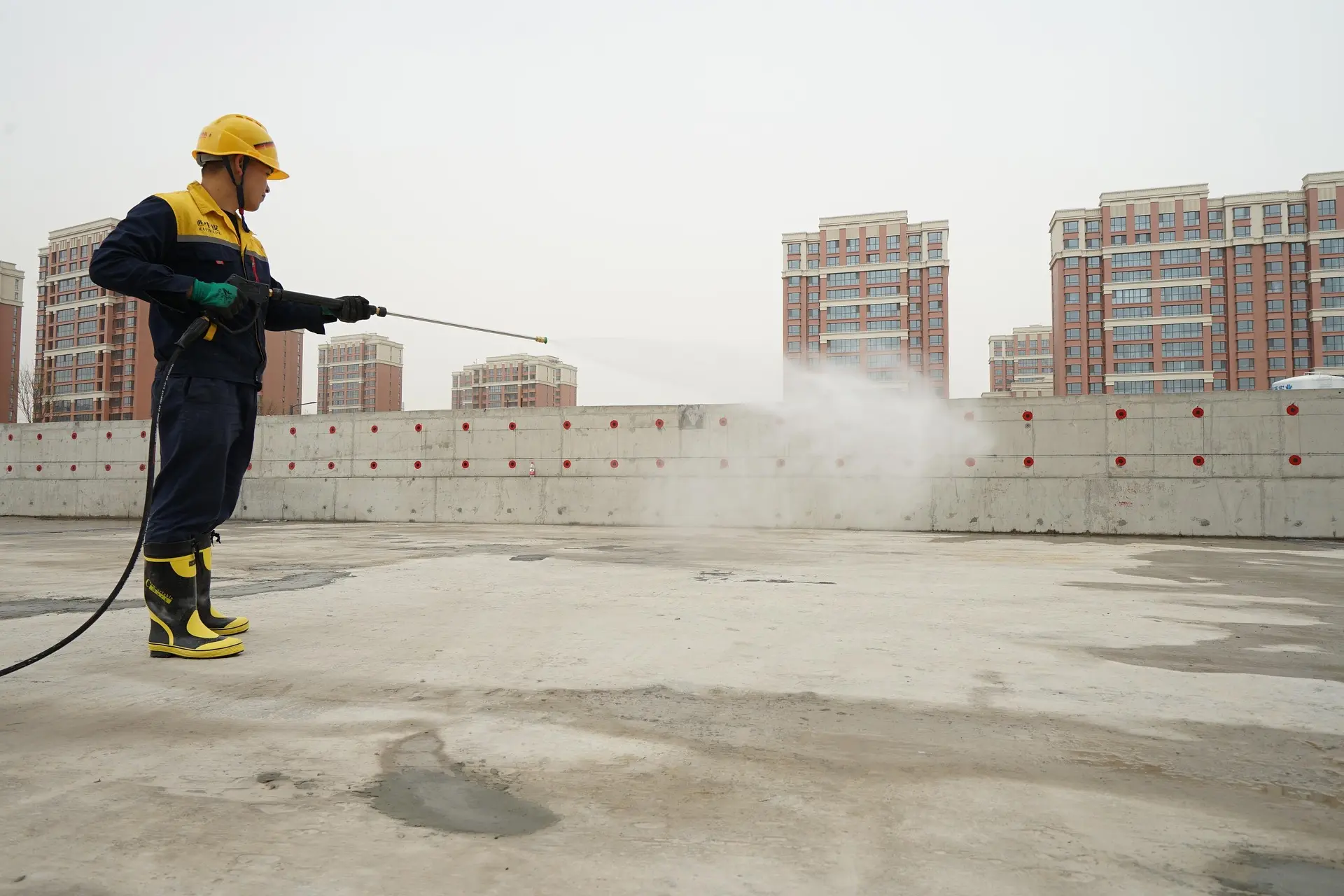 Arbeiter in gelber Schutzkleidung verwendet Hochdruckreiniger auf einer Baustelle mit Betonoberfläche.