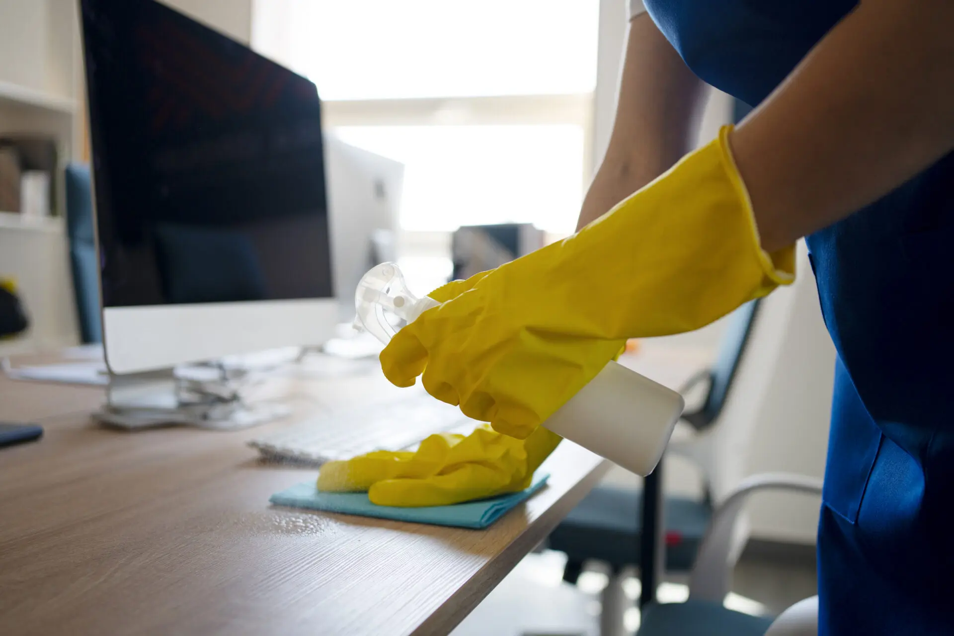 Person mit gelben Gummihandschuhen, die einen Schreibtisch mit einem Reinigungsmittel und einem Tuch reinigt.