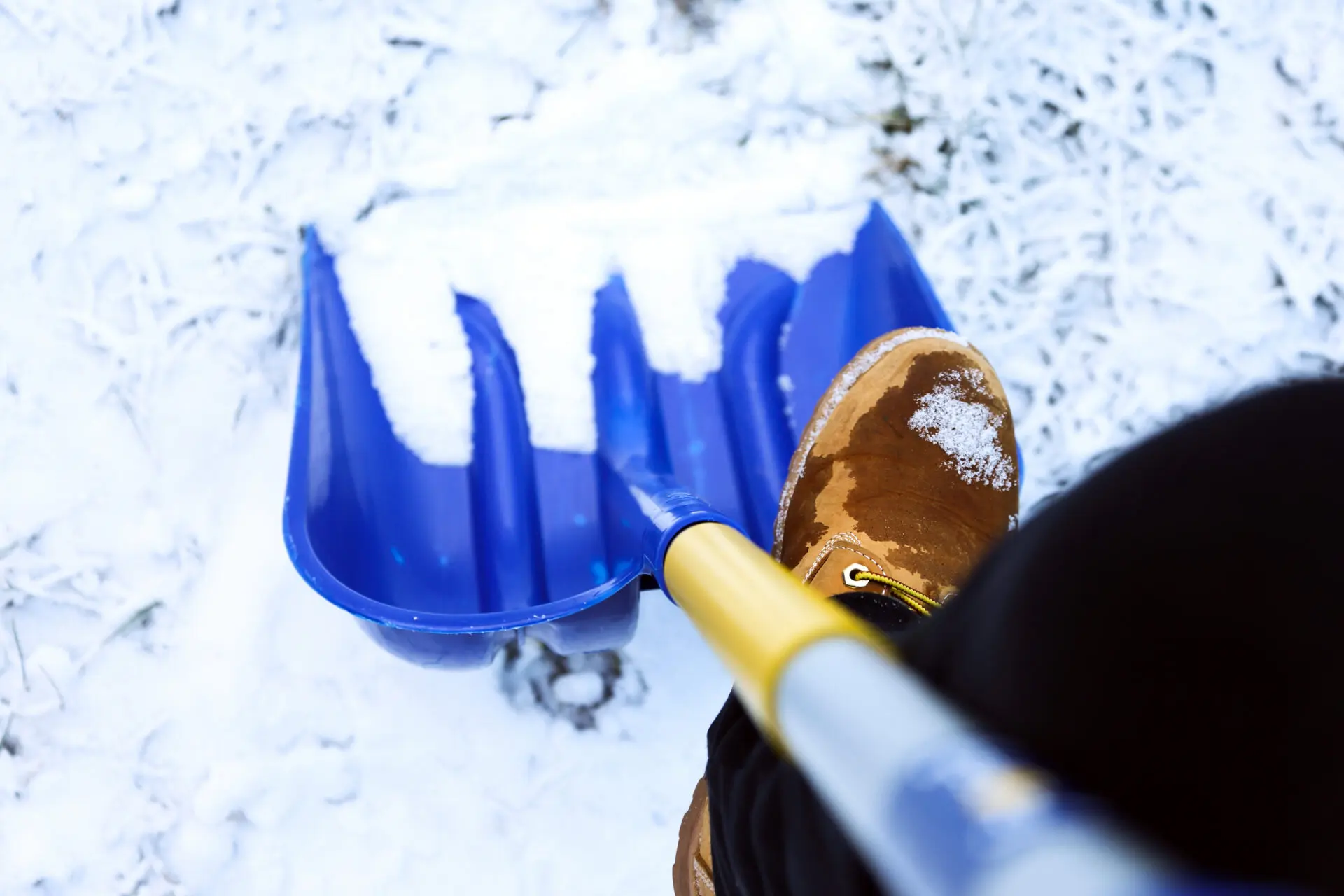 Blaue Schneeschaufel mit Schnee auf dem Schaufelblatt und einem Stiefel im Schnee.